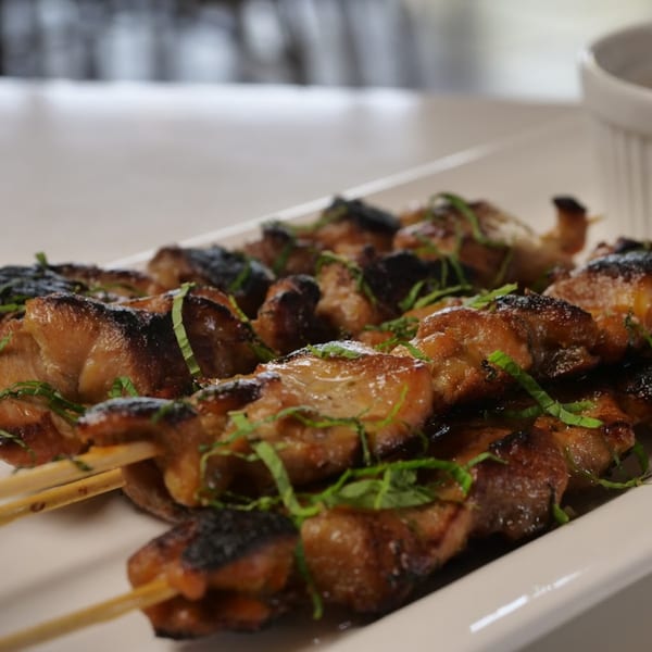Close-up of Vietnamese grilled chicken skewers with charred edges and fresh green herbs on a white plate.
